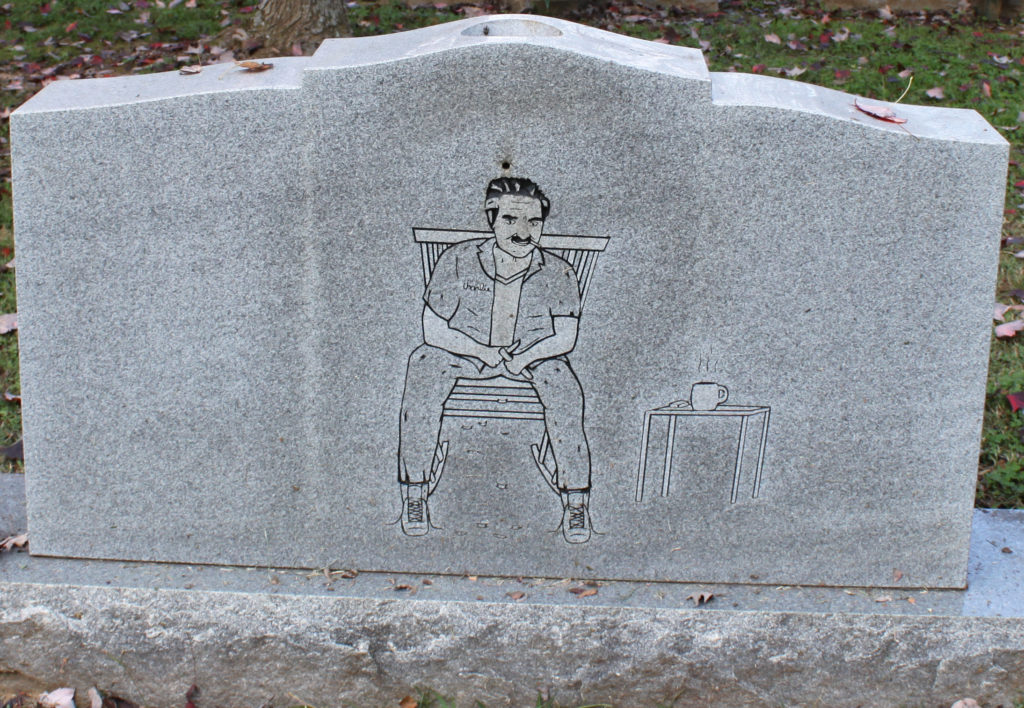 Gravestone depicting a man sitting in a rocking chair, with a steaming cup of coffee, untied shoes, a lit cigarette, all the while he is whittling a piece of wood.